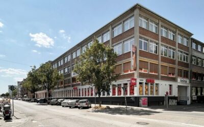 Bild_Felina_Areal Das Felina‑Areal im Mannheimer Stadtteil Neckarstadt Ost: ein großes, mehrstöckiges Gewerbegebäude an einer Straßenecke mit Backstein- und Betonfassade. Reihen großer Fenster prägen die Außenansicht, im Erdgeschoss befinden sich Geschäfte mit roten Schildern. Mehrere Autos parken entlang der Straße, Bäume säumen den Gehweg, der Himmel ist leicht bewölkt.