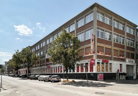 Das Felina‑Areal im Mannheimer Stadtteil Neckarstadt Ost: ein großes, mehrstöckiges Gewerbegebäude an einer Straßenecke mit Backstein- und Betonfassade. Reihen großer Fenster prägen die Außenansicht, im Erdgeschoss befinden sich Geschäfte mit roten Schildern. Mehrere Autos parken entlang der Straße, Bäume säumen den Gehweg, der Himmel ist leicht bewölkt.