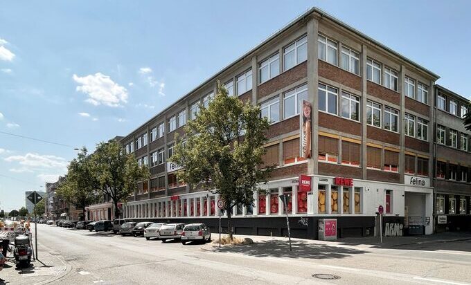 Das Felina‑Areal im Mannheimer Stadtteil Neckarstadt Ost: ein großes, mehrstöckiges Gewerbegebäude an einer Straßenecke mit Backstein- und Betonfassade. Reihen großer Fenster prägen die Außenansicht, im Erdgeschoss befinden sich Geschäfte mit roten Schildern. Mehrere Autos parken entlang der Straße, Bäume säumen den Gehweg, der Himmel ist leicht bewölkt.